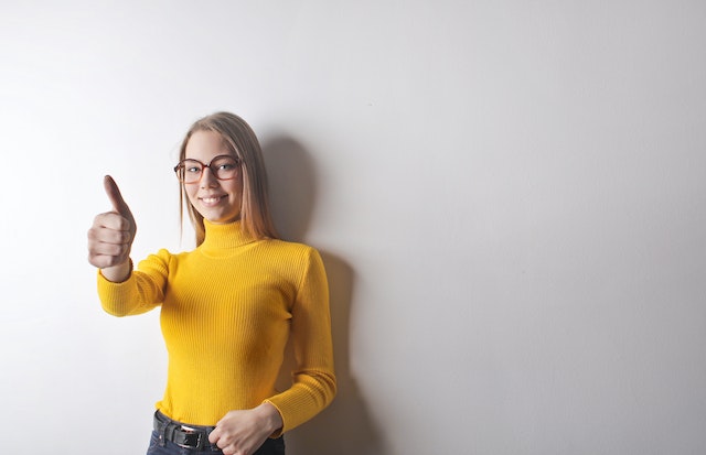 A happy blonde woman in a yellow turtle neck showing thumbs up.